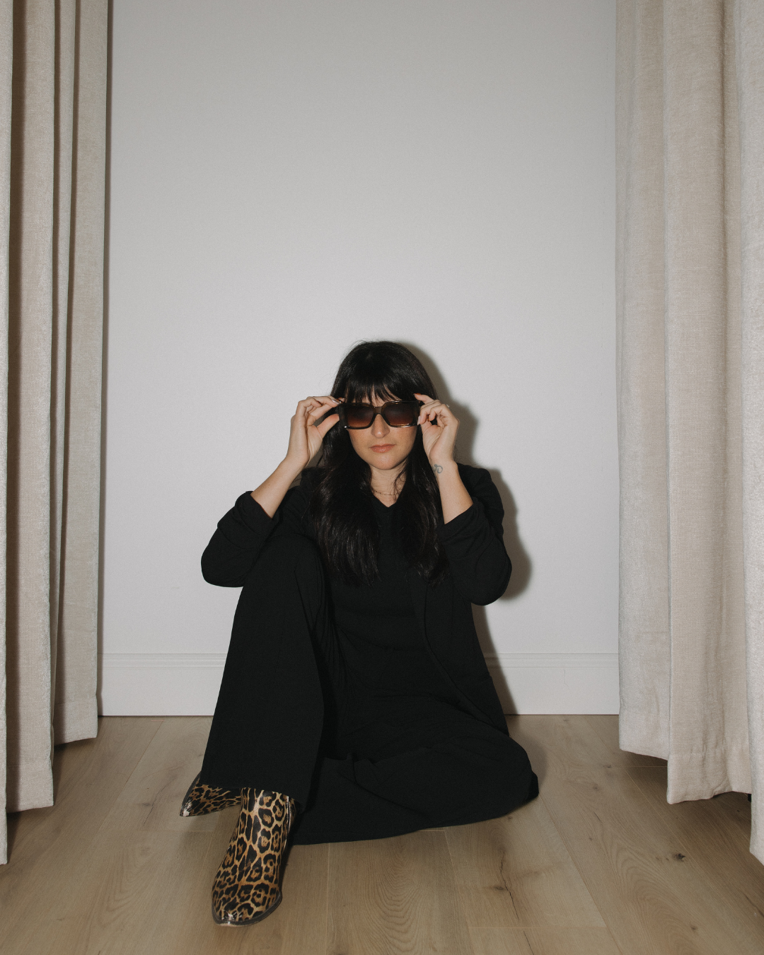 Woman dressed in black sitting on wood floor, adjusting dark sunglasses, with beige curtains on either side.
