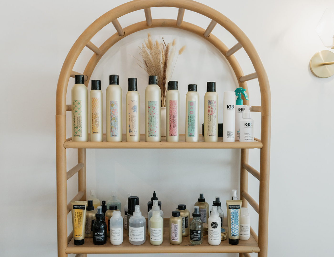 A wooden shelf with three levels holding various bottles of skincare or haircare products, with some large bottles on top and smaller bottles on the lower shelves, against a white wall with a decorative plant in the background.