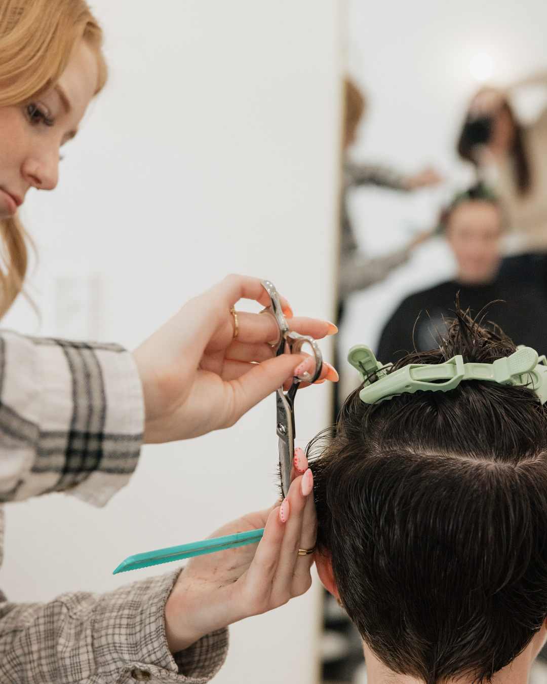 A hairstylist is cutting a person's hair with scissors in a salon. The person has dark hair held with green hair clips, and there are other people in the background, some getting their hair done.