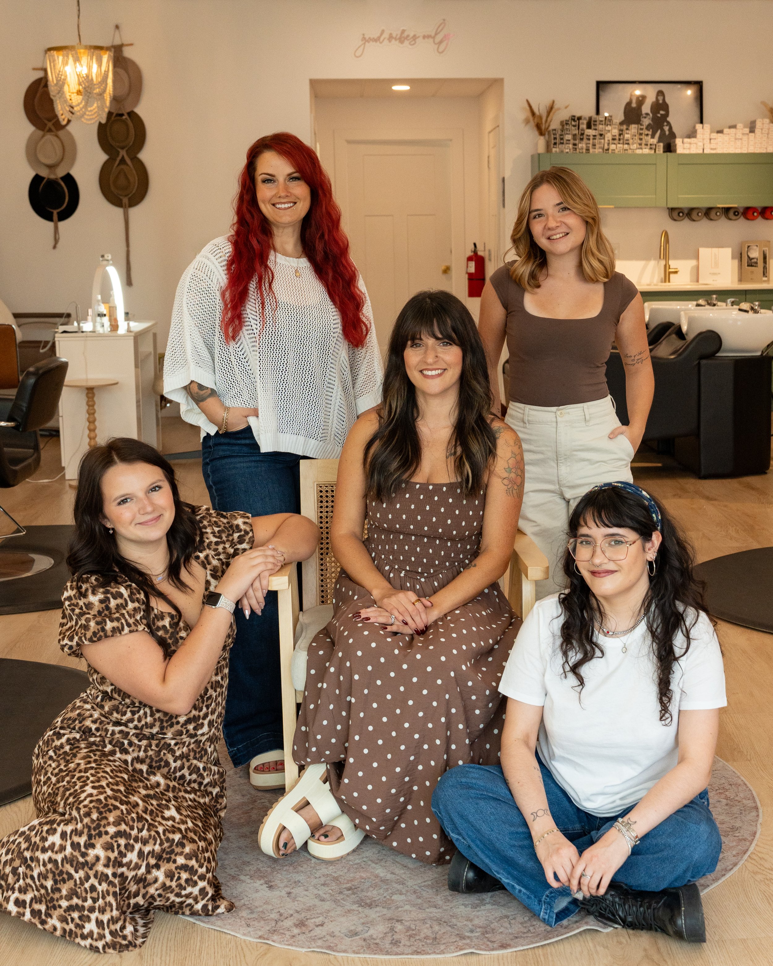 Five women from a salon team pose together inside a bright, welcoming hair salon, smiling at the camera. Two women are seated in front while three stand behind them, surrounded by salon chairs, styling stations, and shampoo bowls.
