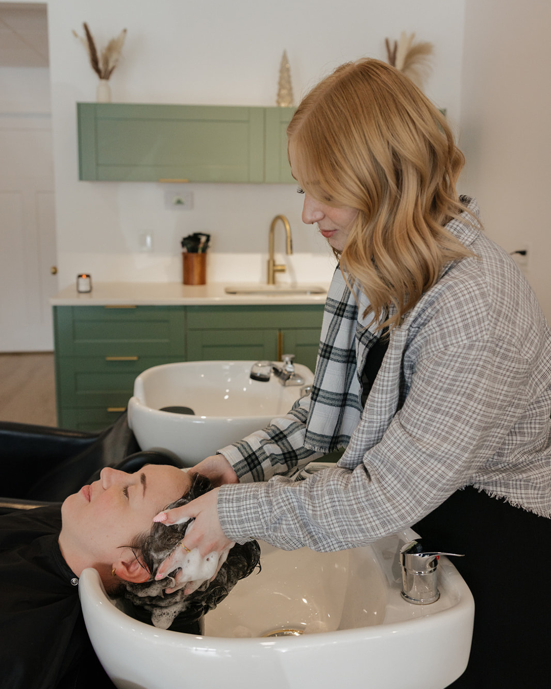 A woman washing her hair in a salon sink while a hairdresser gently rinses it.