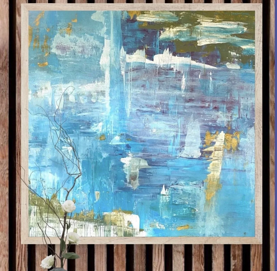 An abstract painting with shades of blue, white, gold, and hints of purple, framed and mounted on a wooden wall with decorative white flowers and twigs in the foreground.