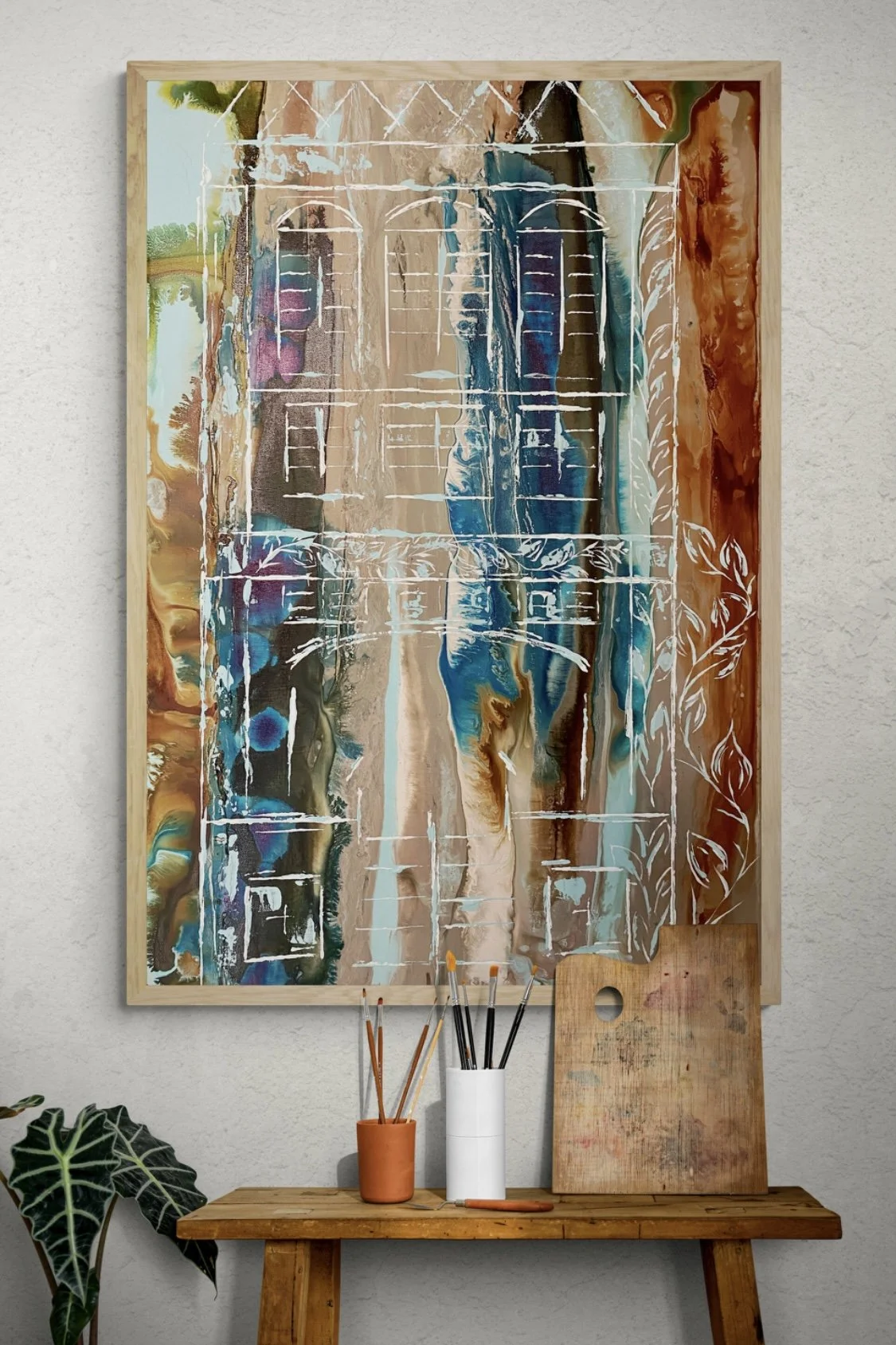 An abstract painting with vibrant, flowing colors including browns, blues, greens, and purples, framed in wood and hanging on a white wall, with art supplies like brushes, a palette, and a wooden board on a wooden table beneath it.