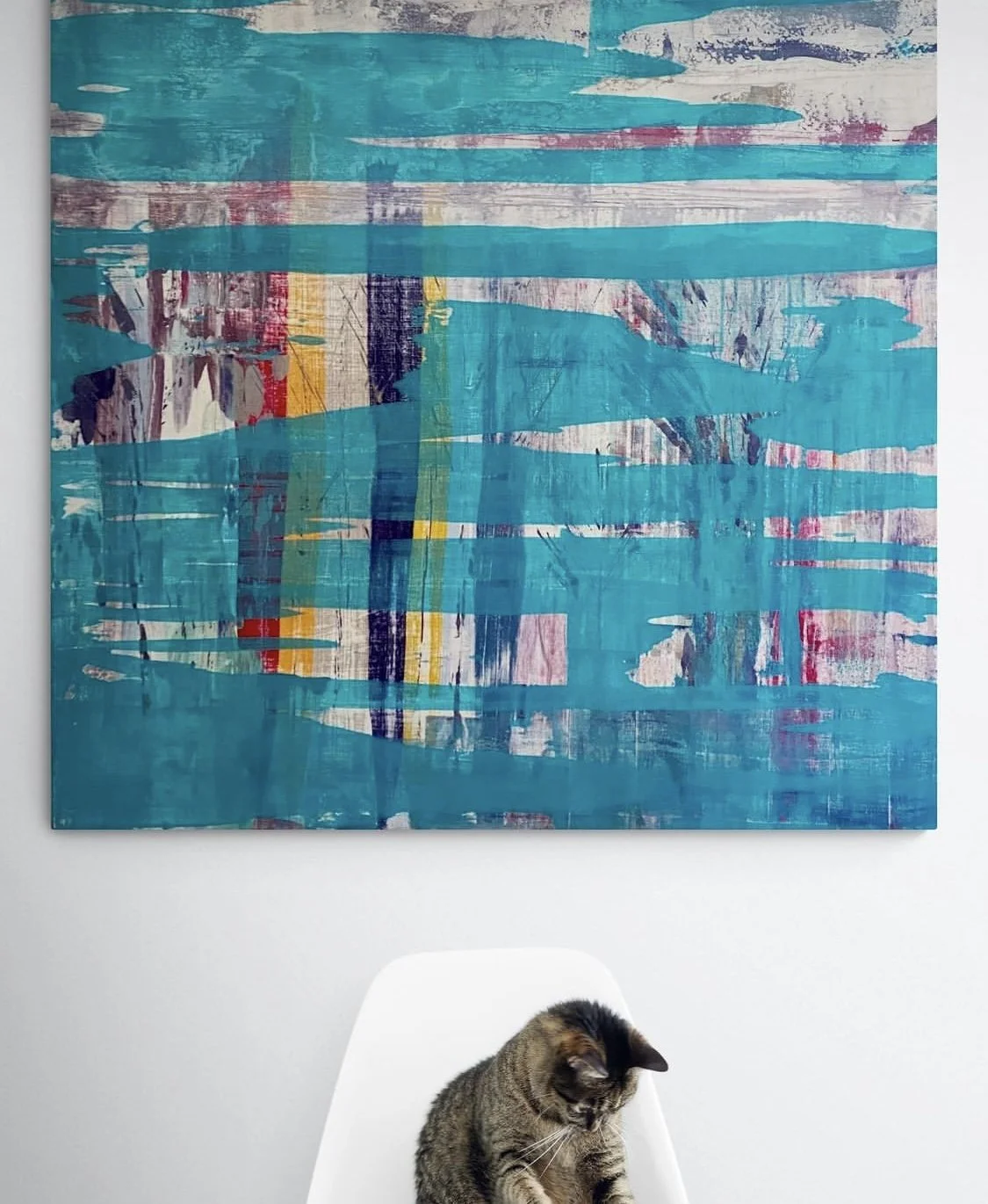 A tabby cat sitting on a white chair below an abstract painting with blue, yellow, red, and purple streaks on a white wall.