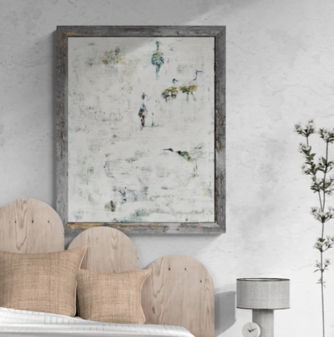 A bedroom with a wooden bed frame, tan pillows, a gray bedside lamp, a small tree, and an abstract painting with a rustic frame on a white wall.