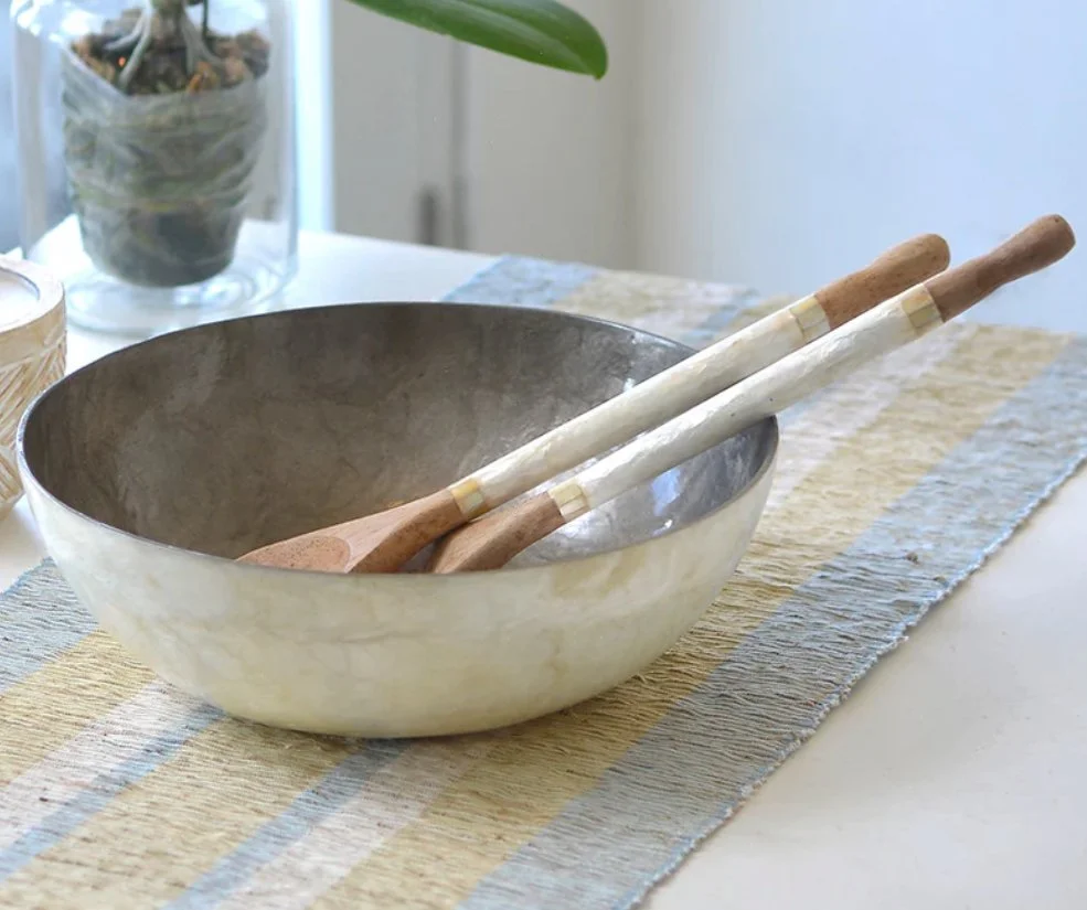 An ethically crafted capiz bowl with two wooden cooking utensils, on a woven table runner in a bright room.