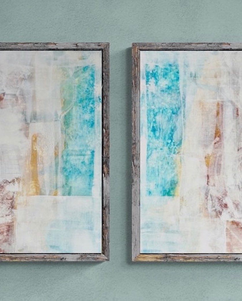 Two abstract paintings with shades of blue, beige, and yellow, framed in rustic wood, hanging on a light green wall.