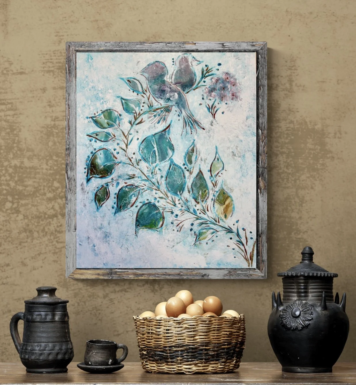 Decorative still life with a basket of eggs, two black ceramic jars, a small black cup and saucer, set on a wooden table in front of a textured beige wall and a framed watercolor painting of blue and green leaves and birds.