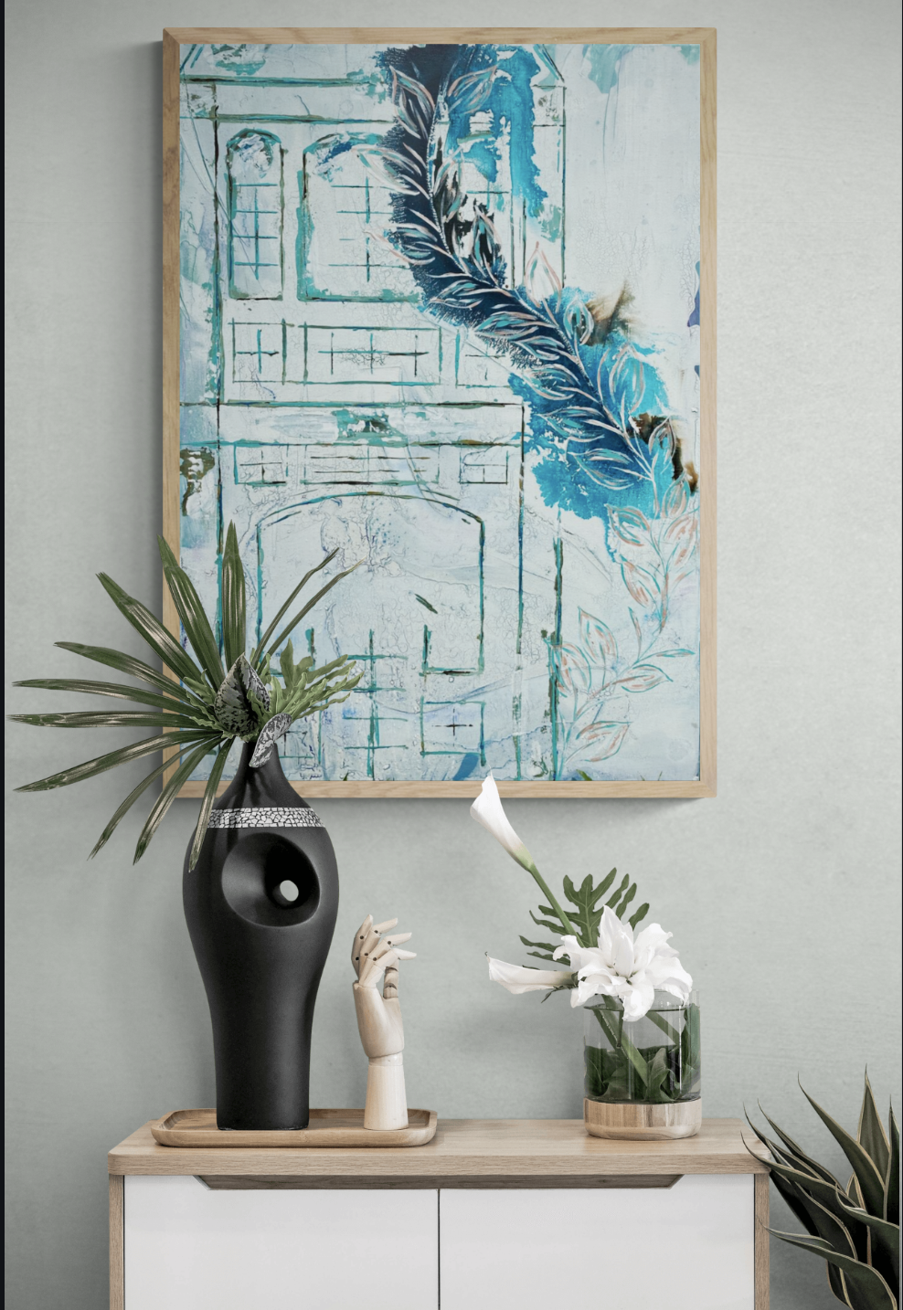 Modern interior decor with a wooden-framed abstract painting in blue and white shades, a black vase with green tropical leaves, and a small glass vase with white flowers on a light wood sideboard against a light green wall.