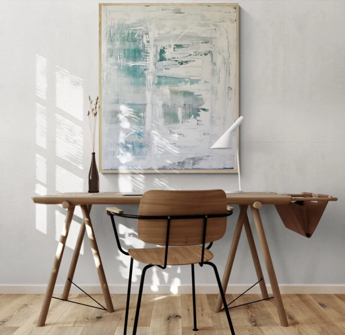 A minimalist workspace with a wooden desk, a matching wooden chair, a white desk lamp, a tall brown vase with dried grasses, and abstract artwork on the wall, with sunlight creating patterns on the wall.
