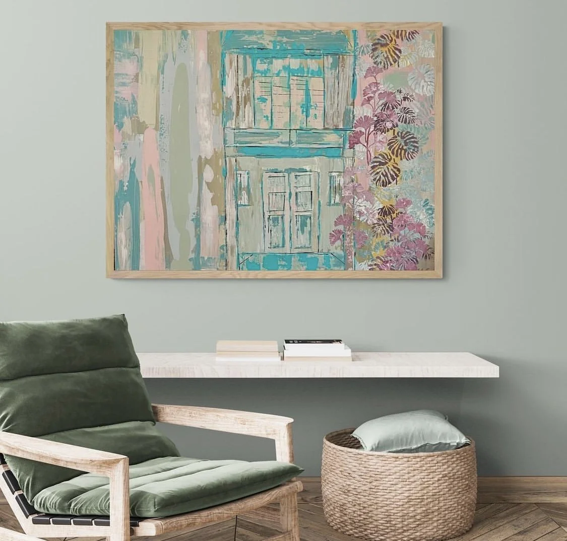 A living room with a large abstract painting of a house and trees hangs on a mint green wall. A white floating shelf holds books, a green cushioned armchair with wooden armrests is in the foreground, and a woven basket with a cushion is on the wooden