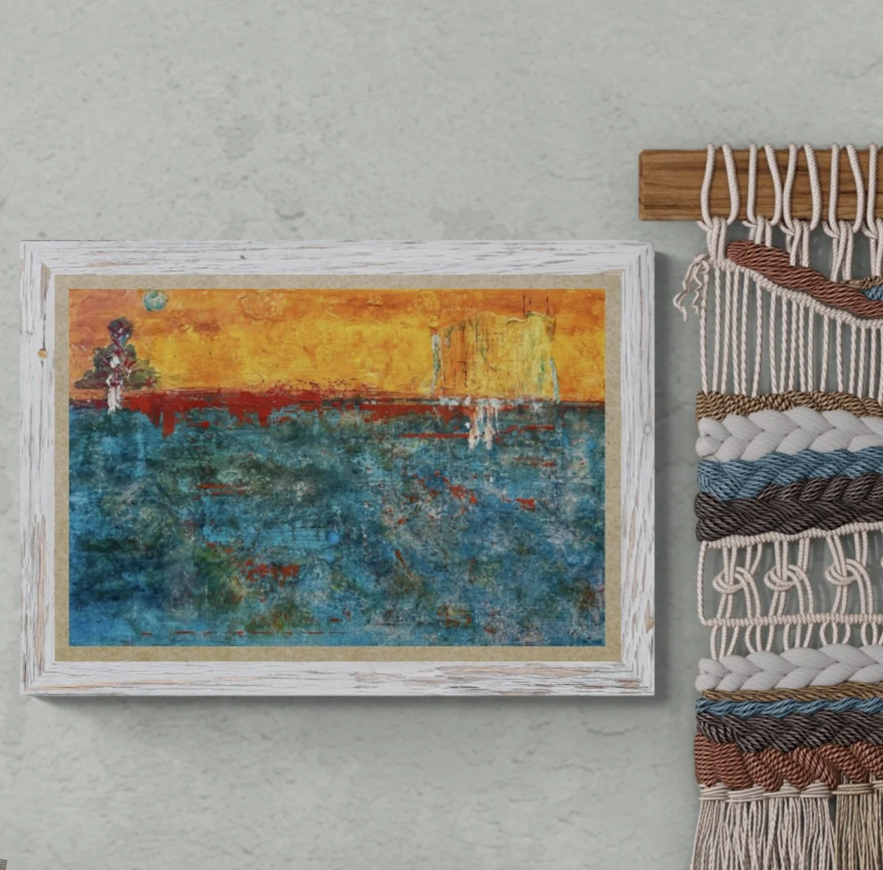 Abstract painting of a sunset with water, a tree on the left, and a building or structure on the right, with a star or planet visible in the sky, framed in white and hung on a wall next to woven wall art.