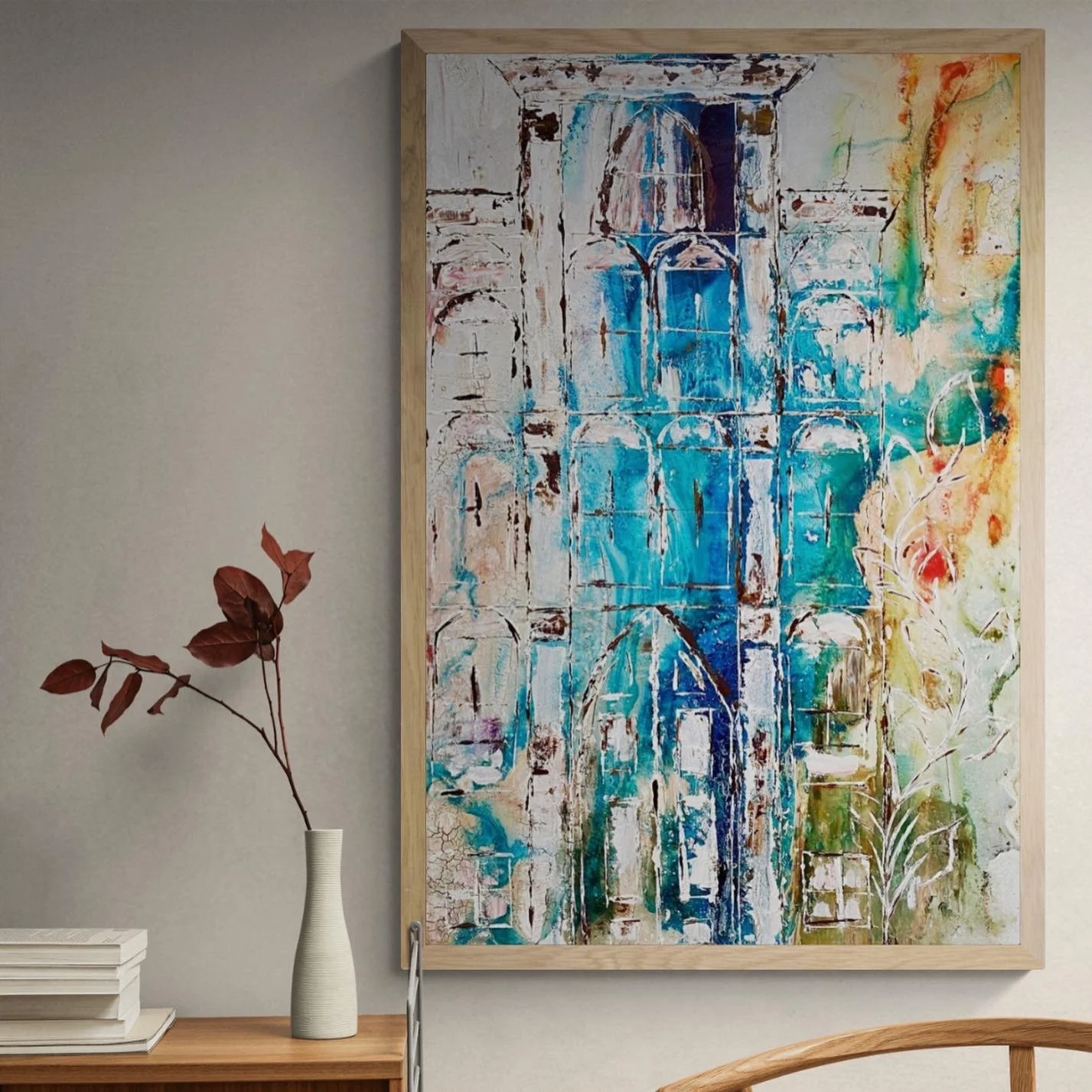 Large abstract painting with blue, white, beige, and orange colors, framed and hung on a plain wall, with a small wooden table below holding a white vase with brownish-red leaves and a few stacked books.