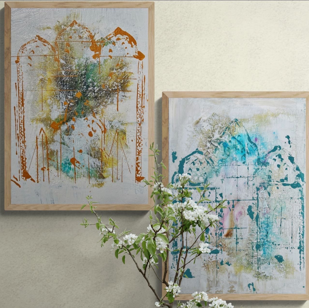 Two abstract paintings with floral elements and a small branch with white flowers in front, set against a beige wall.