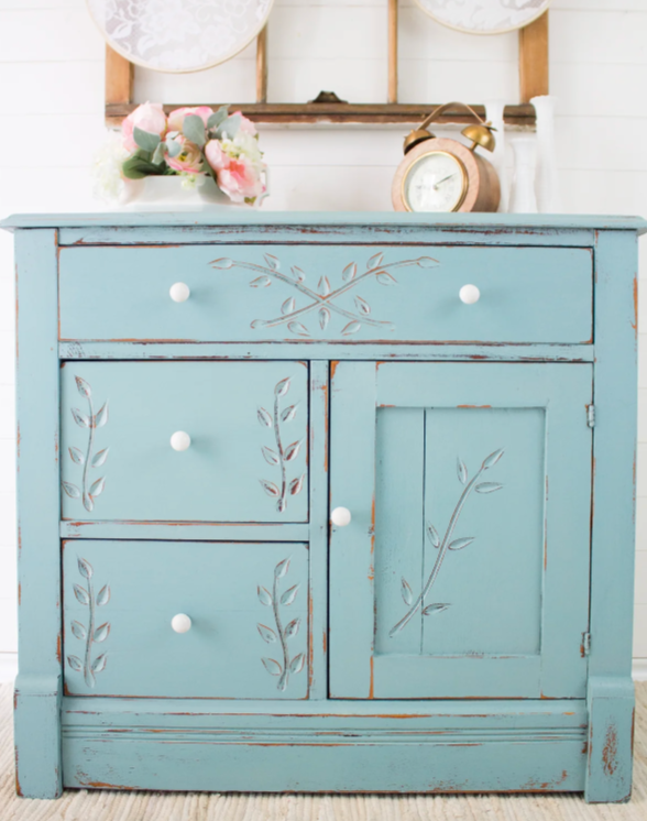 Distressed blue wooden sideboard with white knobs and leaf motifs, topped with a floral arrangement, clock, and wall decor.