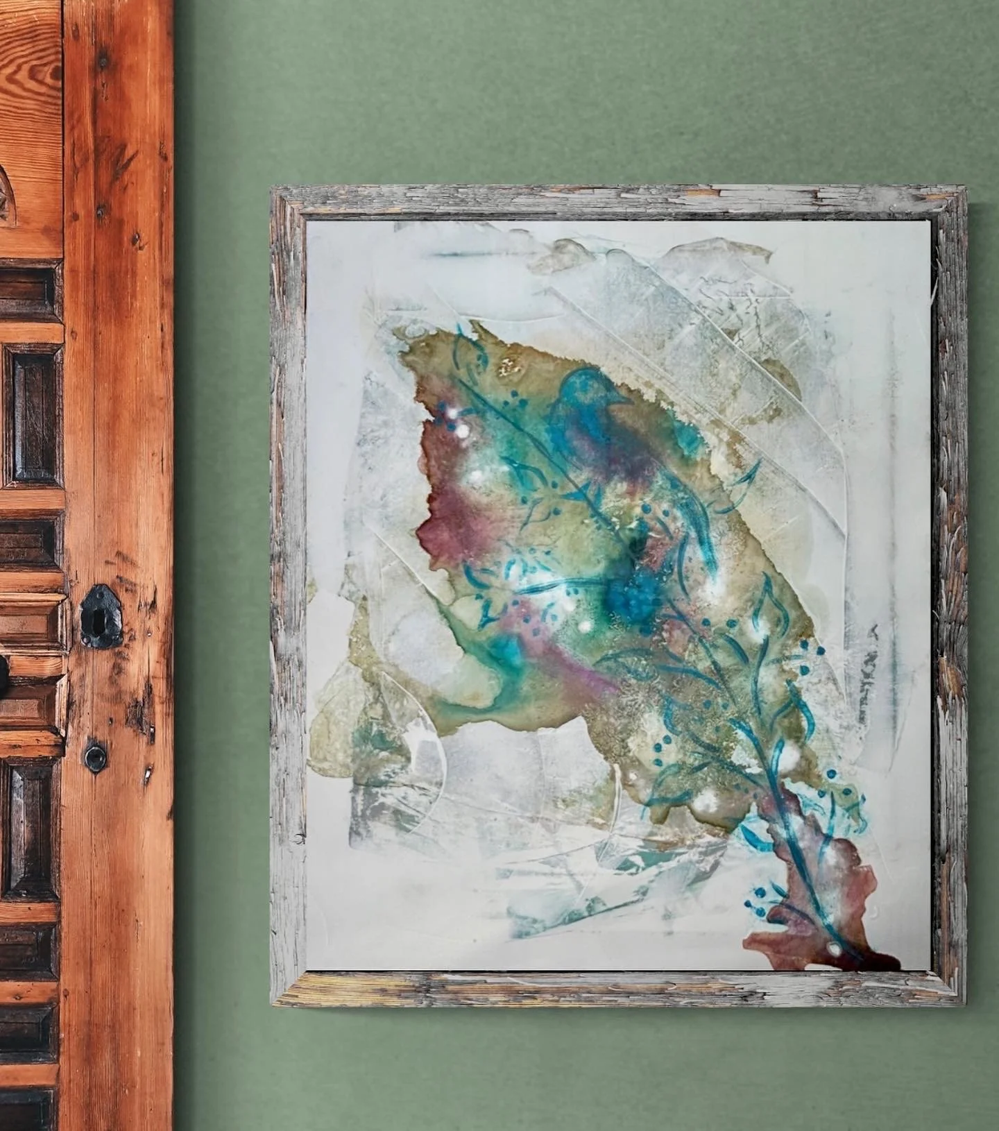 Abstract watercolor painting with shades of green, blue, purple, and brown framed in weathered wood, hanging on a green wall next to a wooden door.