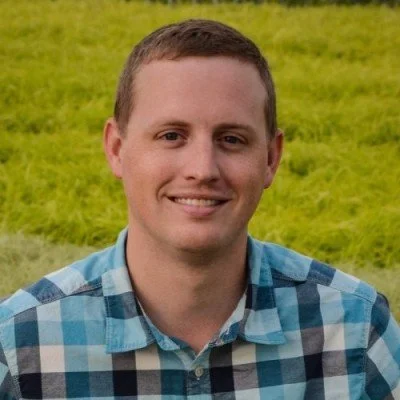 Portrait of a young man with short brown hair, smiling, wearing a blue plaid shirt, standing outdoors in front of a green grassy field.