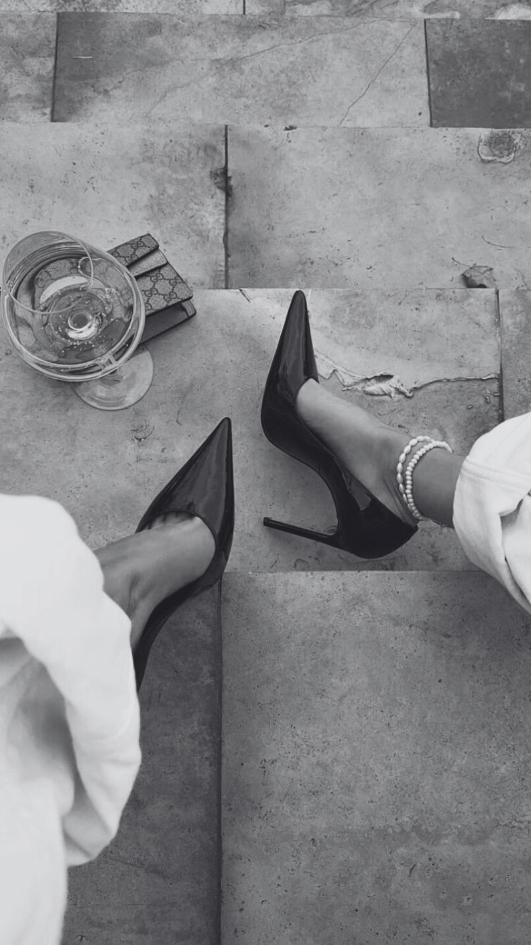 Photo en noir et blanc montrant des jambes de femme avec chaussures à talons hauts, un bracelet de perles, une bouteille de vin, et un portefeuille Gucci, prises depuis une vue aérienne.