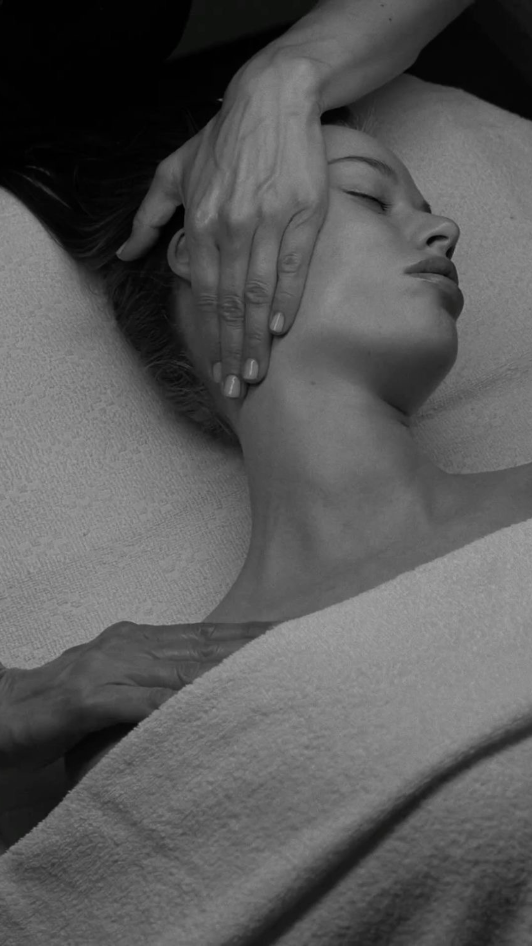 Une femme repose avec les yeux fermés, une main d'une personne âgée touche son visage, dans un style en noir et blanc.