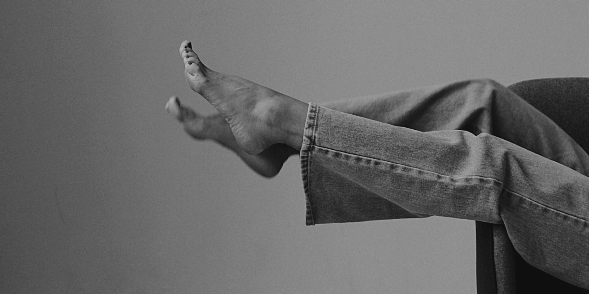 Jambes d'une personne assise, portant un jean, avec les pieds levés dans le vide, en noir et blanc.