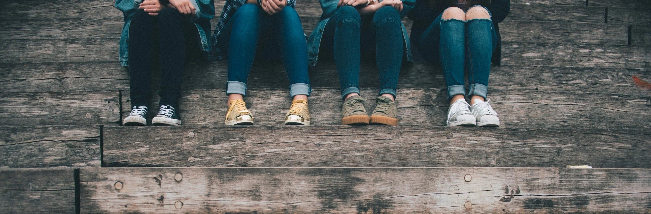 Four teens sitting on wooden steps, showing only their legs and feet. They are wearing casual clothing and sneakers.