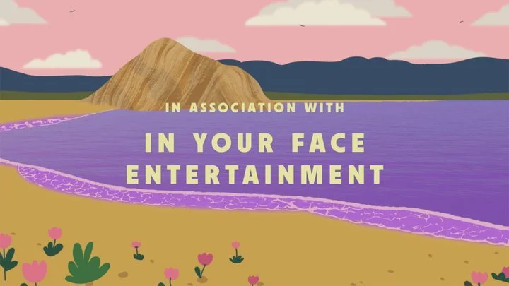 A scenic landscape with a purple lake, a sandy shore with flowers, a large mountain, and a partly cloudy sky. Text over the image reads: In association with In your face entertainment.