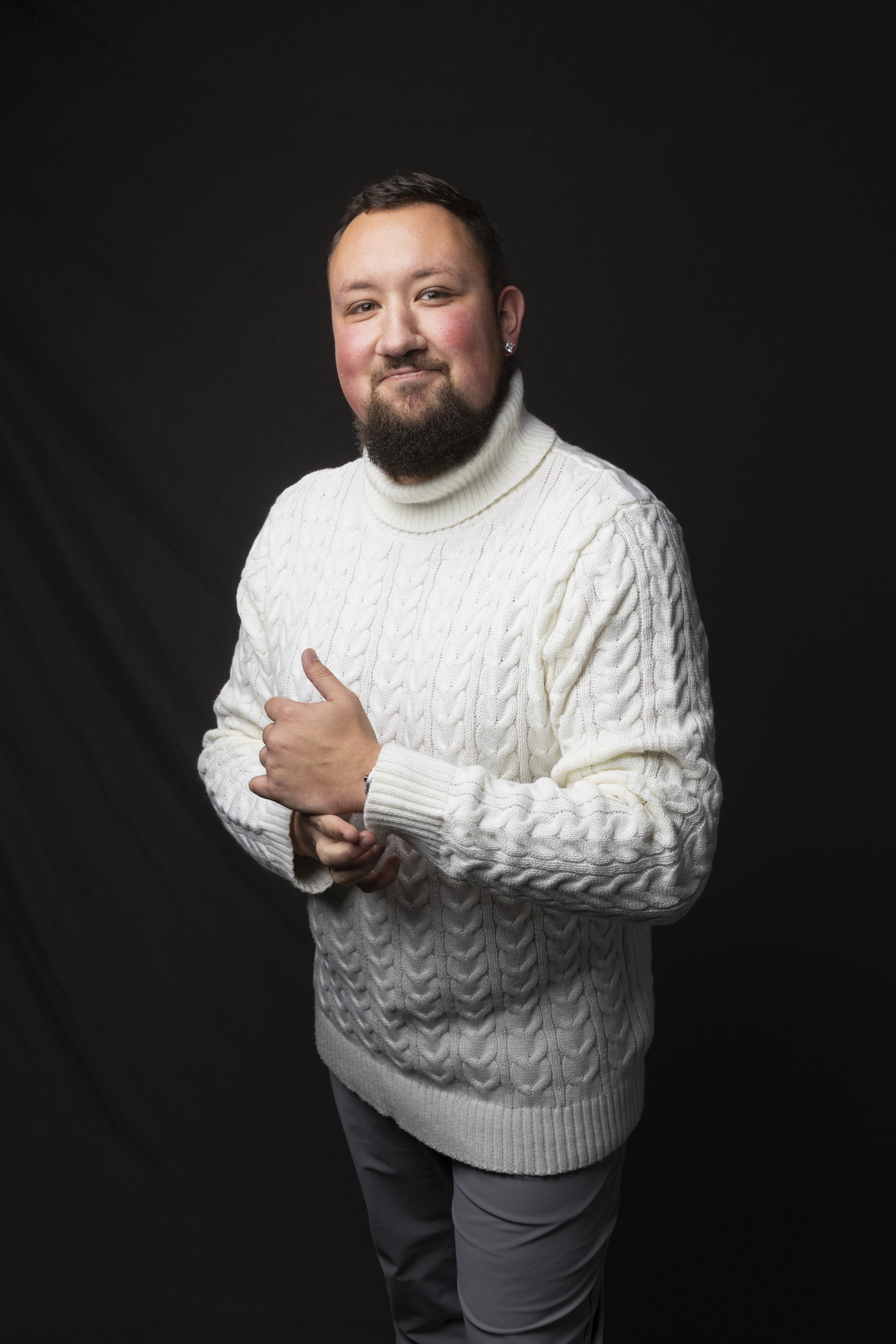 A man with a dark beard and short hair, wearing a white cable-knit turtleneck sweater and gray pants, stands against a black background with a slight smile and his right hand resting on his chest.