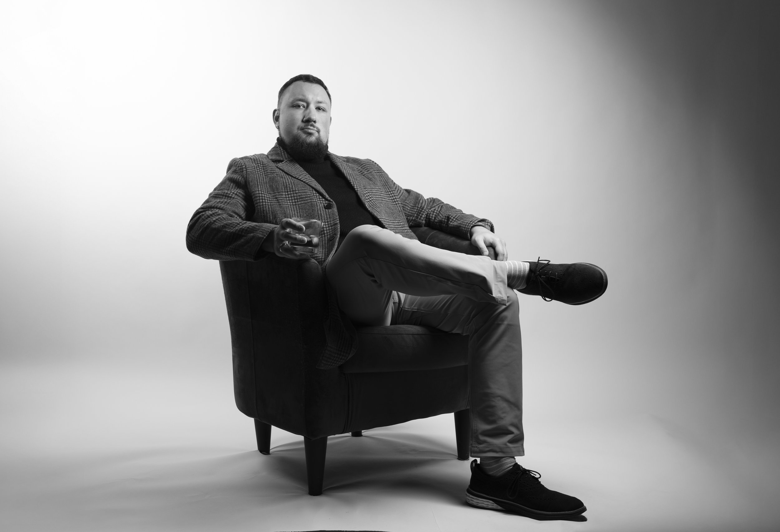 Black and white photo of a man sitting in a chair with one leg crossed over the other, holding a glass, and looking confidently at the camera.