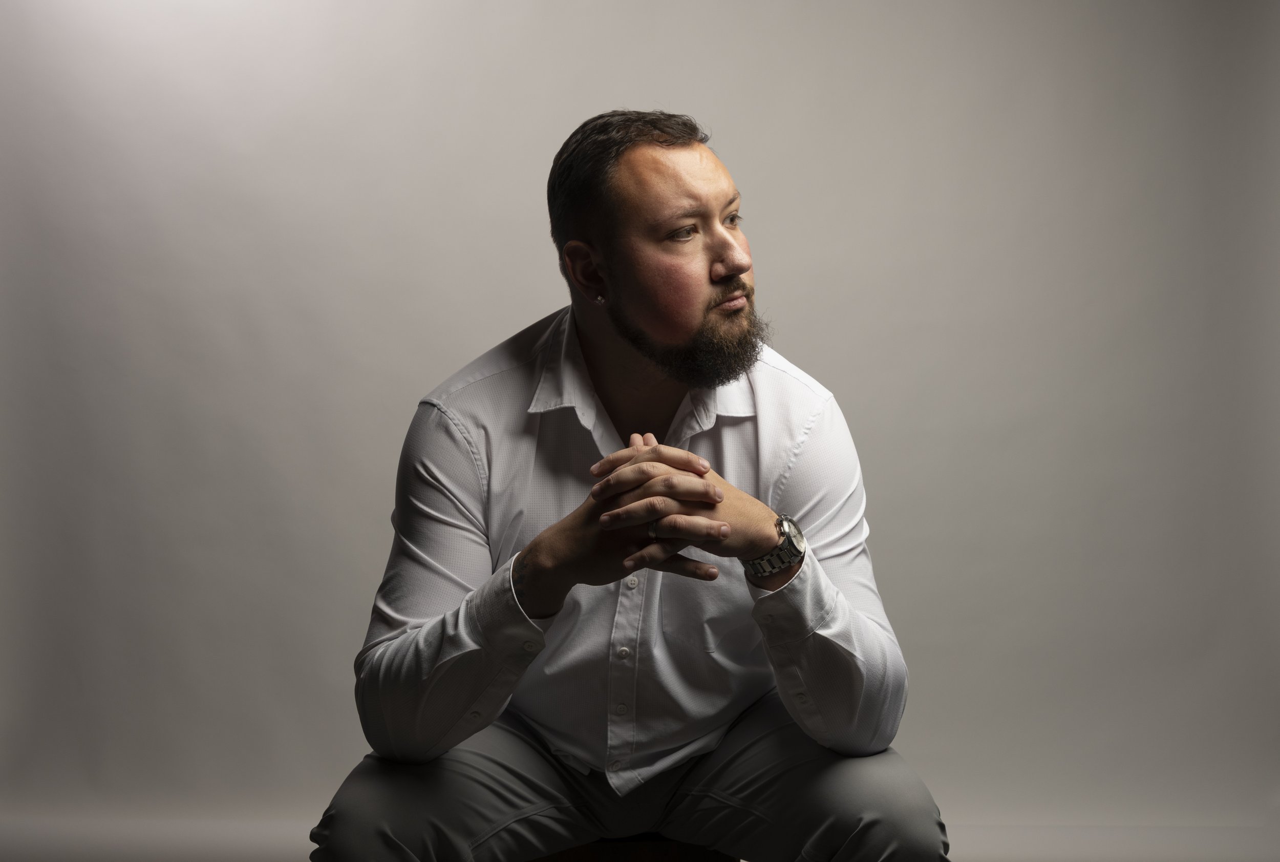 A man with a beard and mustache, wearing a white dress shirt and wristwatch, sits with his hands clasped, looking contemplative against a neutral background.