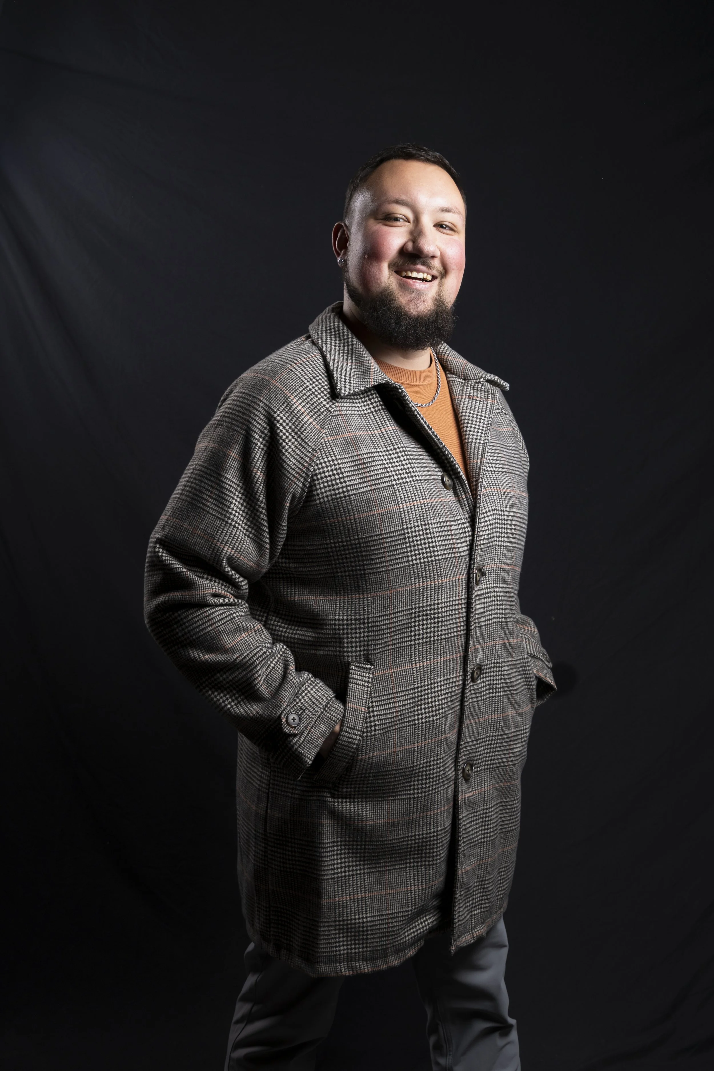 Smiling man with a beard and earrings wearing a plaid coat and an orange shirt, standing against a black background.