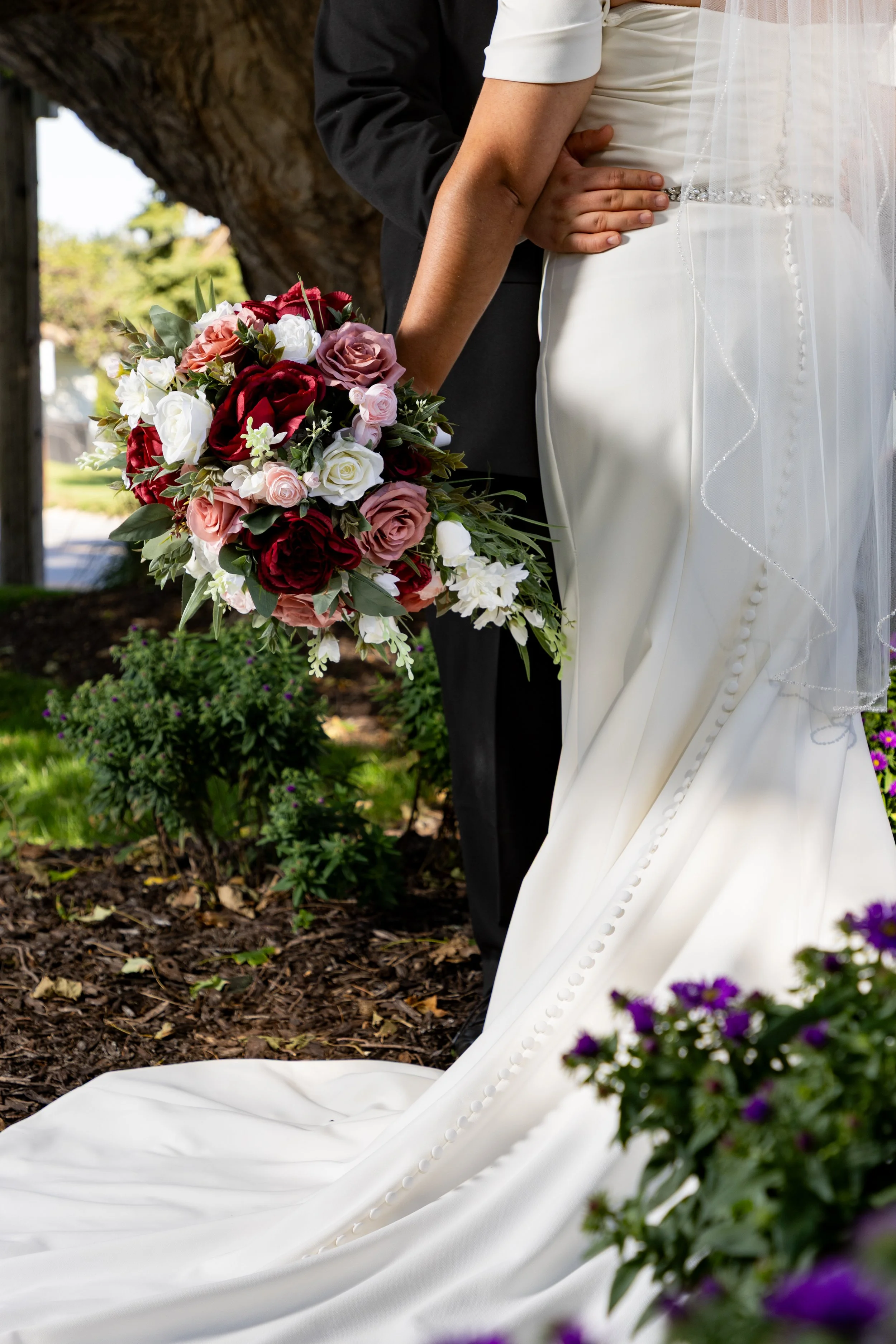 Close-up of a bride and groom holding a bouquet of red, pink, white roses, and other flowers, with the bride wearing a white wedding dress and the groom in a black suit, outdoors near a tree.