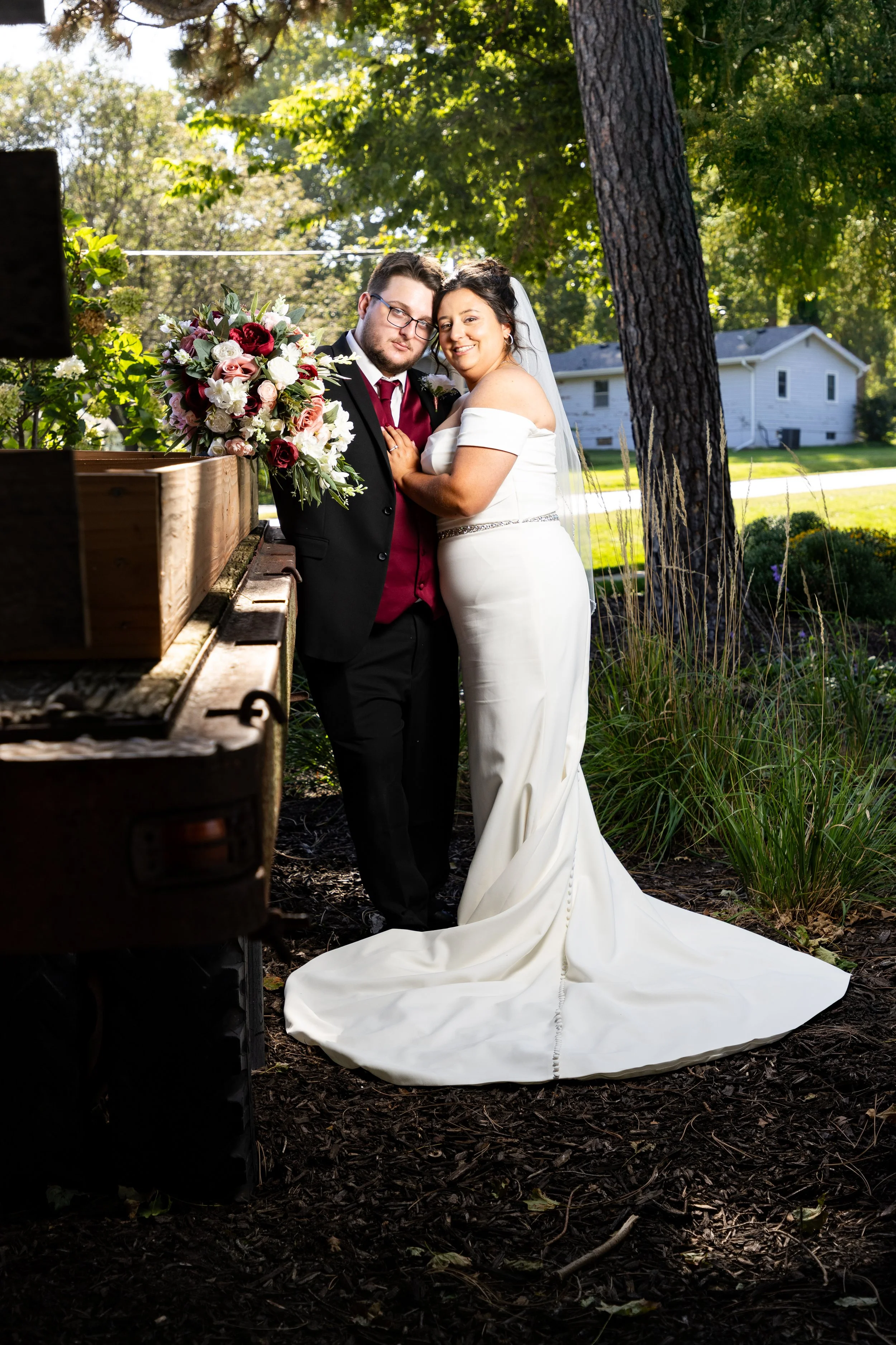 A couple on their wedding day, dressed in formal wedding attire, standing outdoors in a garden with lush greenery and a house in the background. The bride wears a white off-shoulder gown with a train and veil, and the groom wears a black tuxedo with a maroon shirt and vest. They are close together, smiling and embracing, with a large bouquet of flowers on a wooden bench beside them.