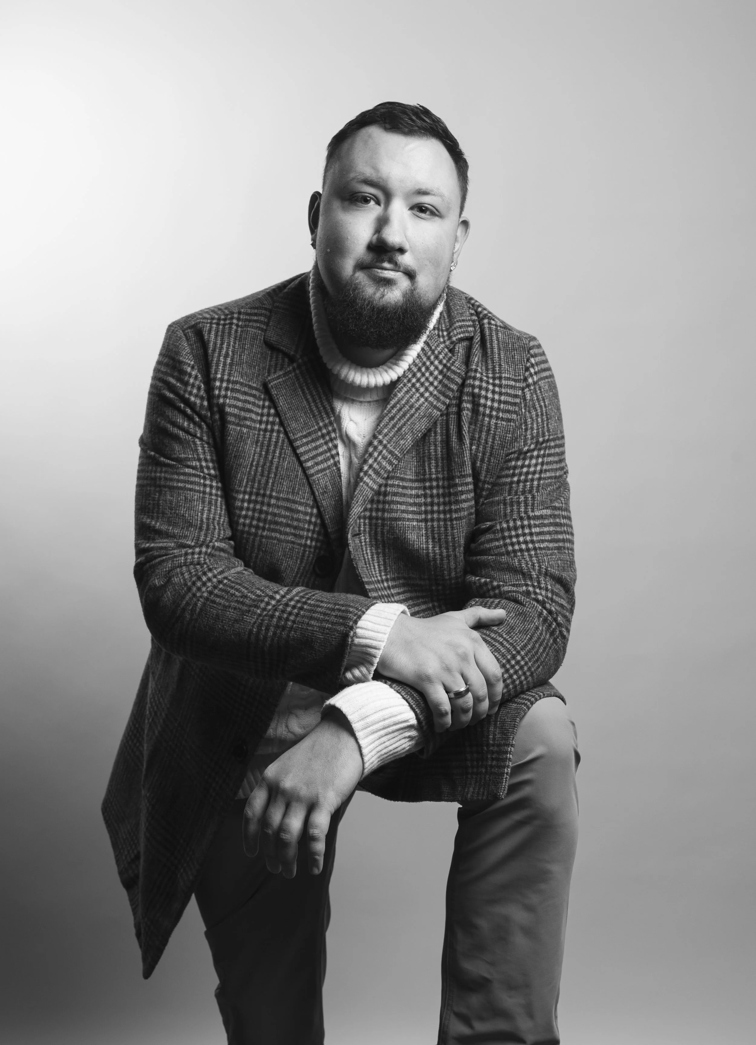 Black and white portrait of a man sitting on a stool, wearing a checkered blazer and a turtleneck sweater, with short hair and a beard, looking directly at the camera.