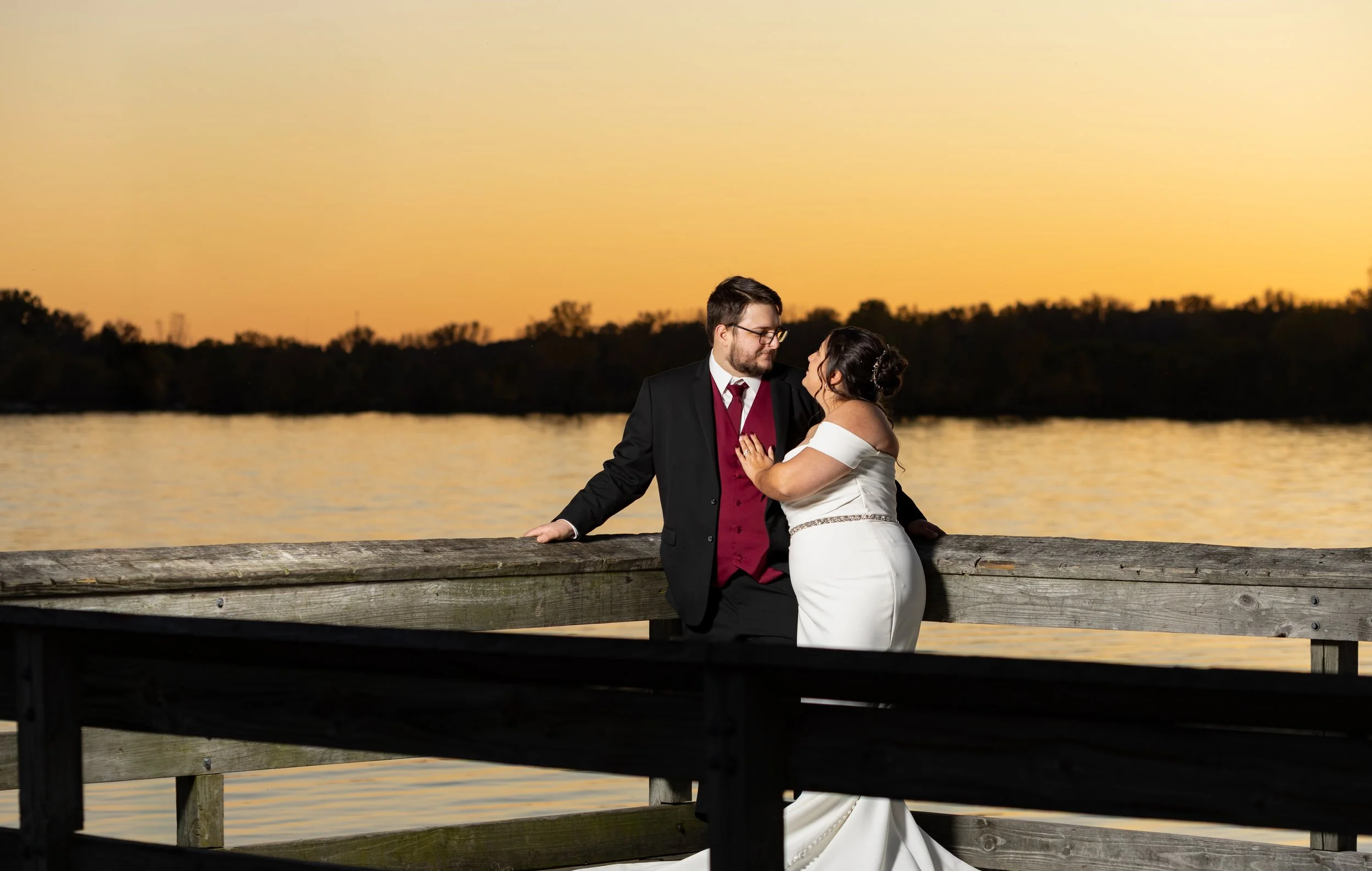 A newlywed couple standing on a wooden dock by a lake during sunset, gazing into each other's eyes.