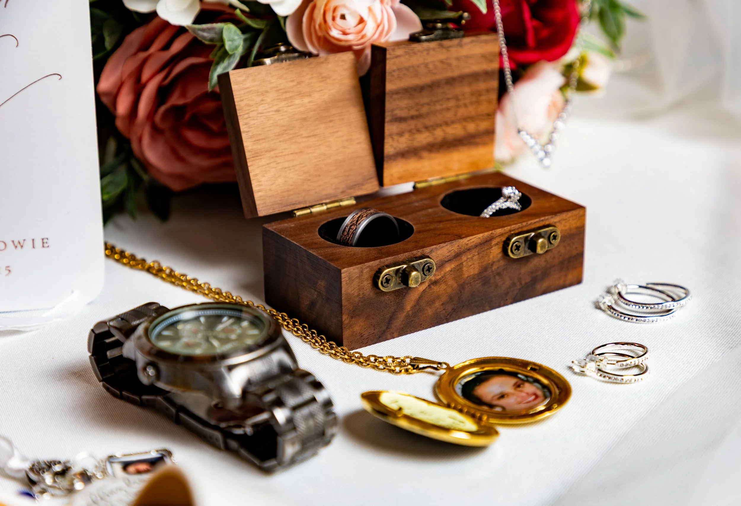 A wooden jewelry box with rings inside, surrounded by watches, necklaces, and earrings on a white surface, with pink and red flowers in the background.