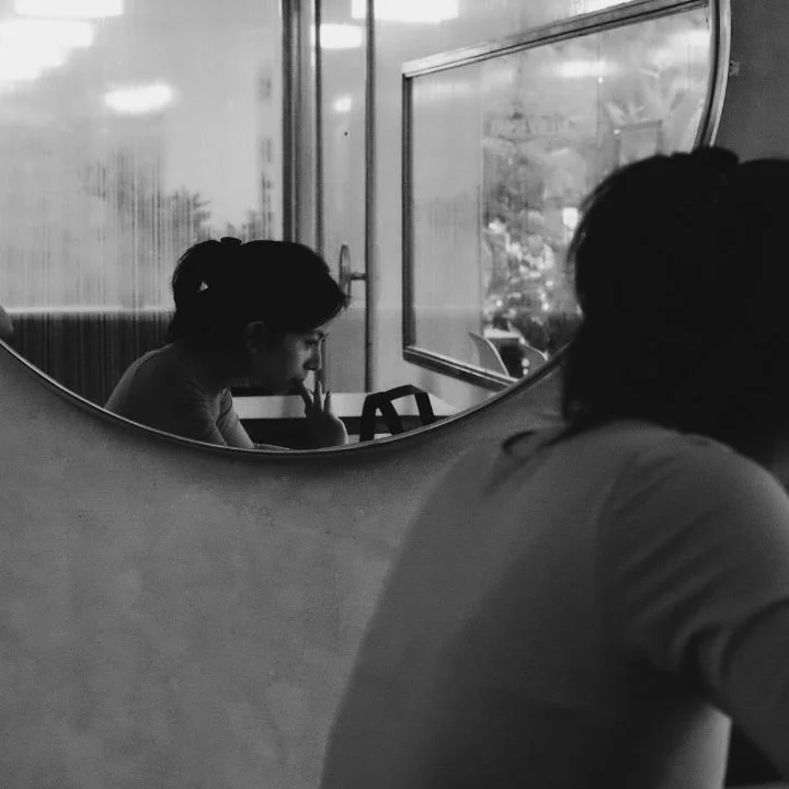 Person seen in mirror sitting quietly by window, appearing deep in thought.