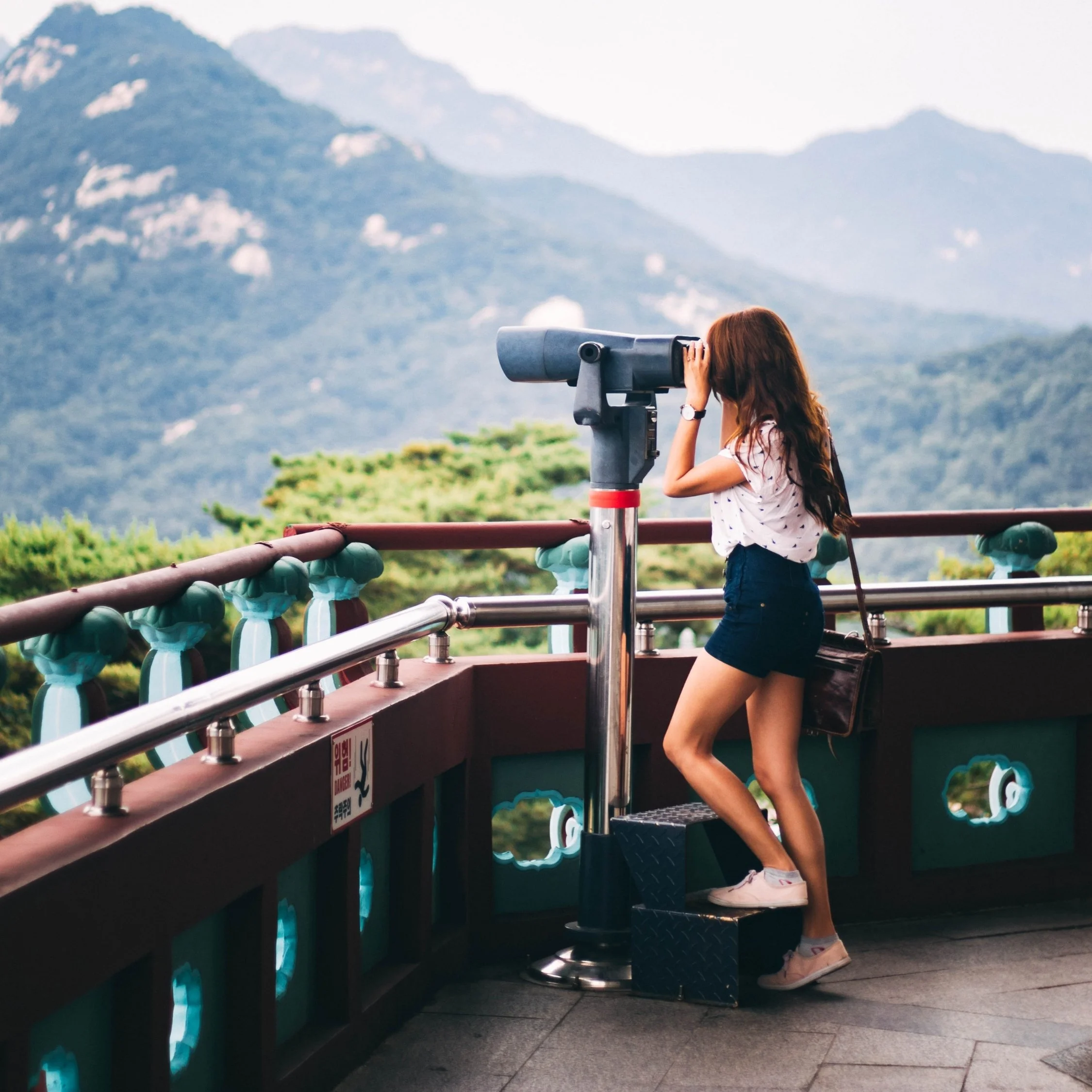 Young woman looking through viewing binoculars at mountain landscape from scenic overlook.