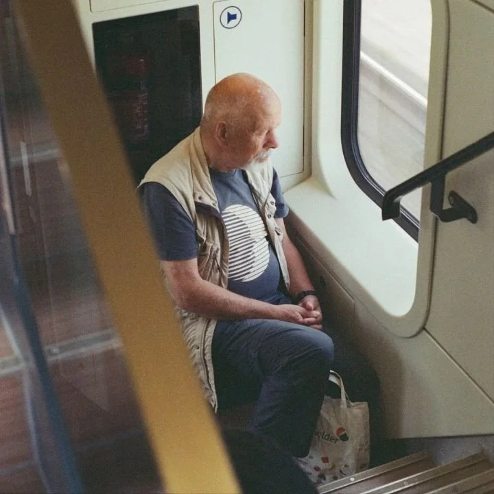 Older man sitting alone on train, gazing out window thoughtfully.