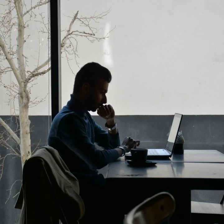 Man sitting at table by window, looking thoughtfully at laptop with coffee nearby.