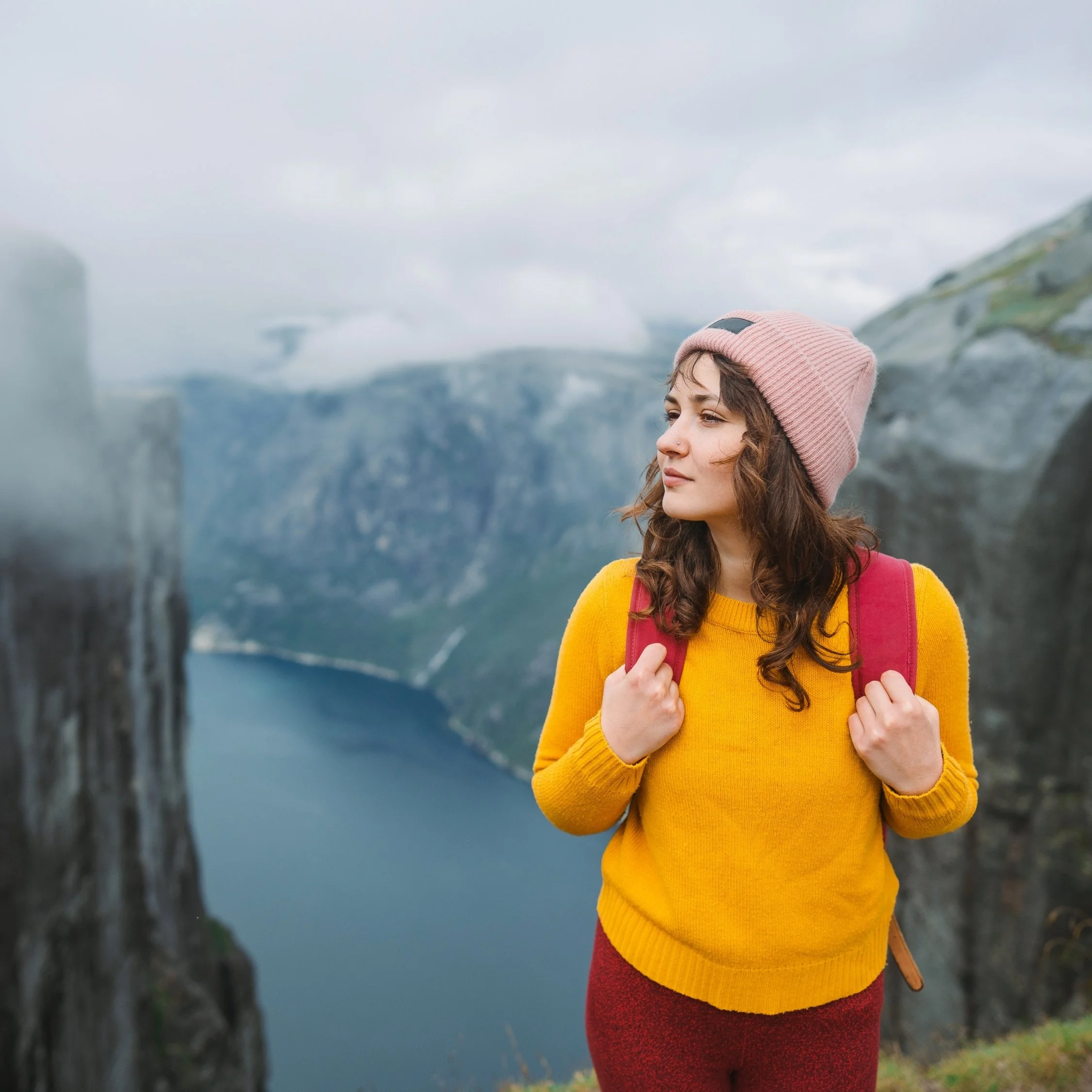 Woman with backpack standing near cliff overlooking fjord.