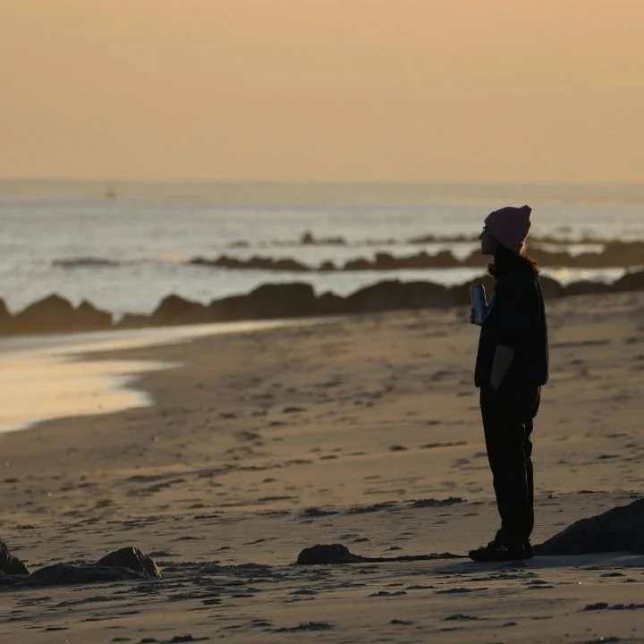 Person standing alone on beach at sunset holding a cup and looking at ocean.
