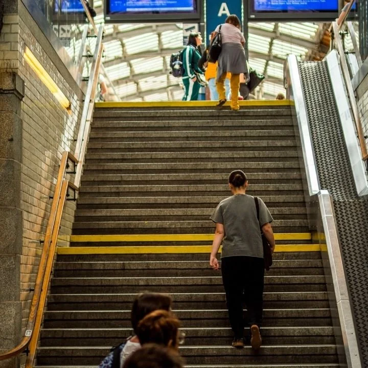 Person walking up wide station stairs toward bright exit with others ahead.