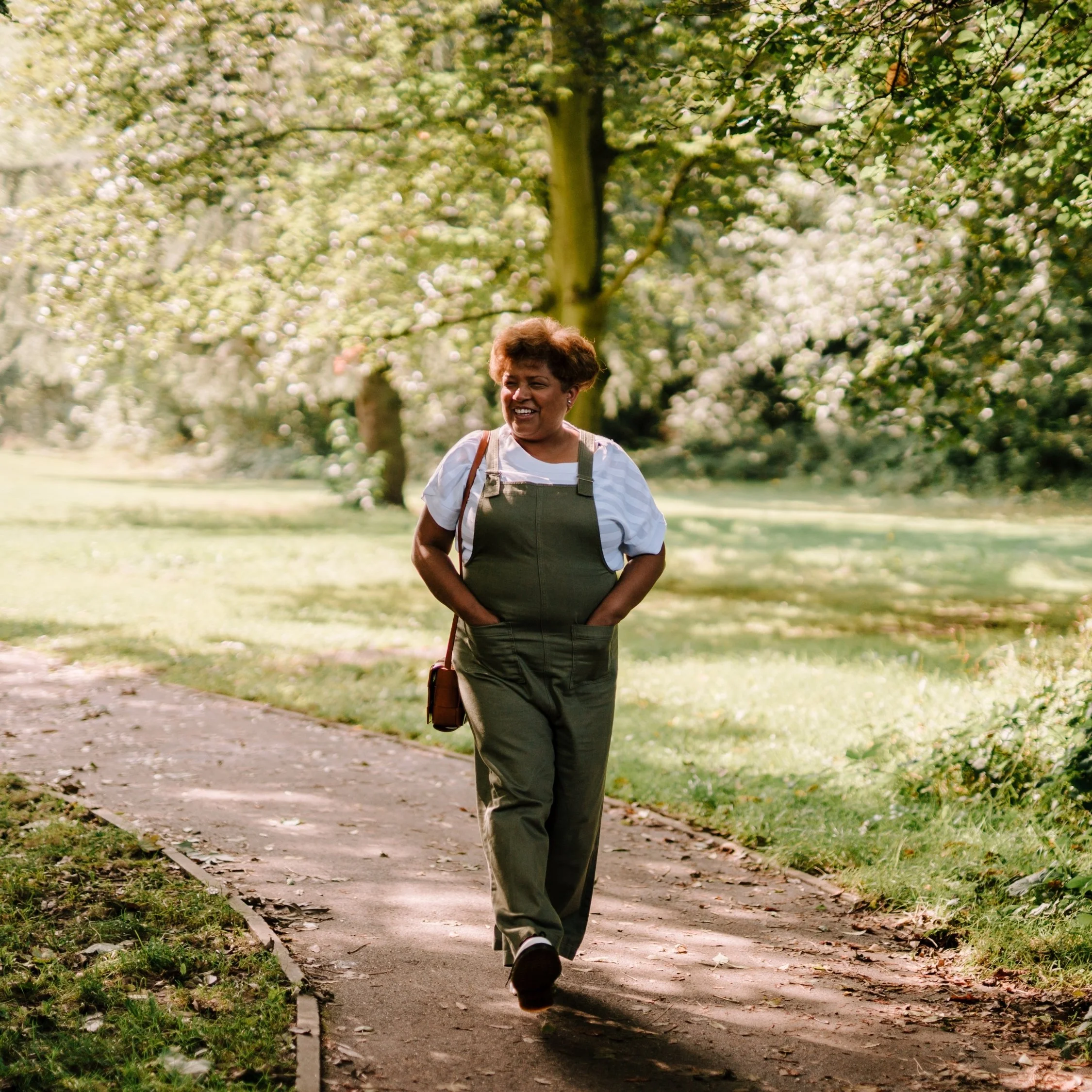 Middle-aged woman smiling while walking along a tree-lined park path on a sunny day.