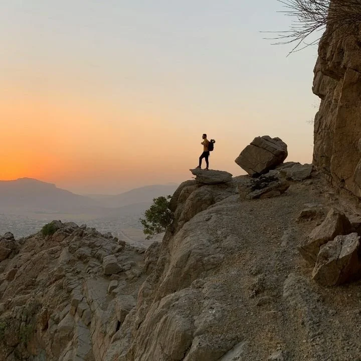 Person with backpack standing on rocky cliff overlooking valley at sunset.