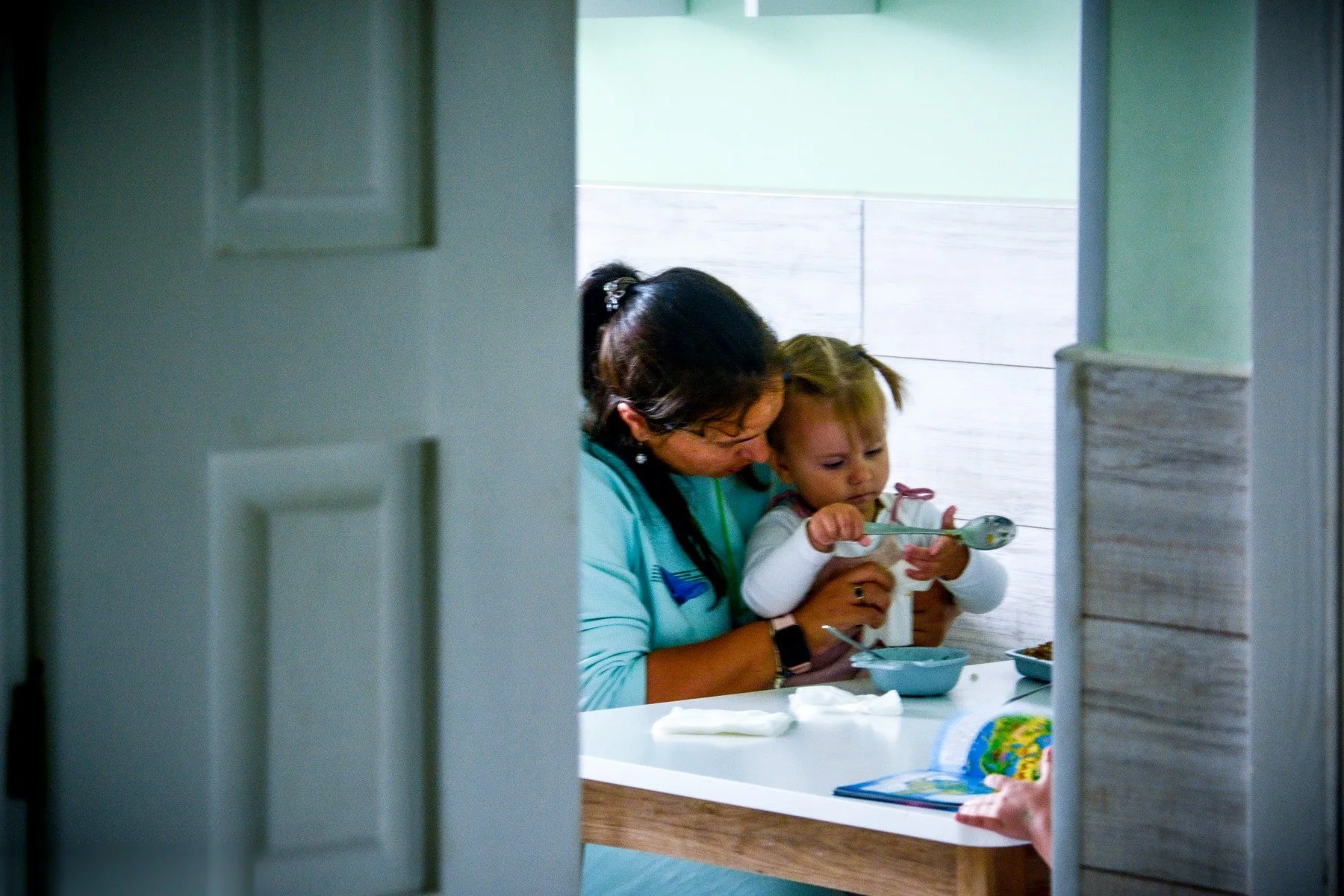A woman holding a toddler at a table, the child reaching for a spoon