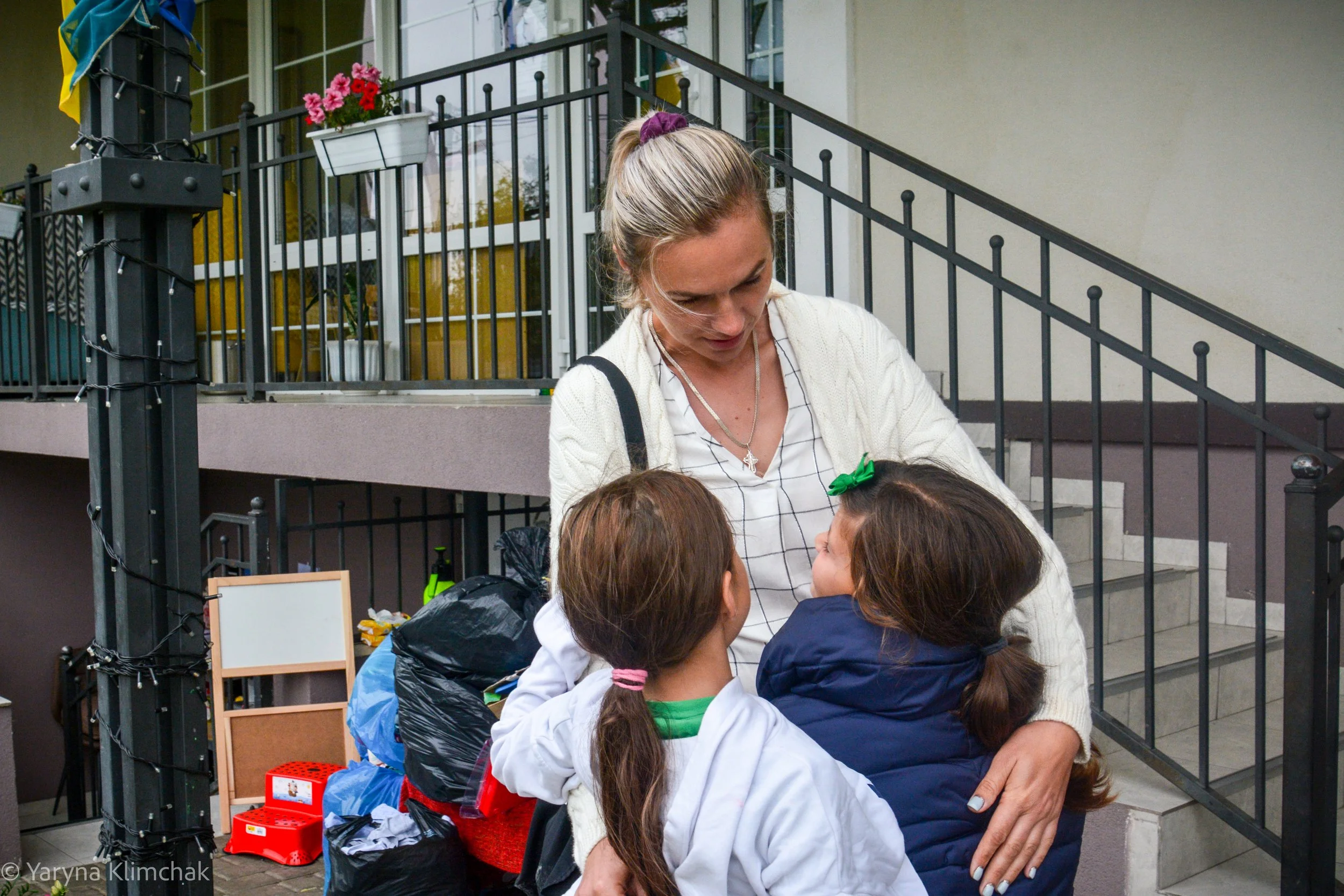 A woman in a white cardigan bends to embrace two girls at the foot of an outdoor staircase at City of Goodness, a Ukrainian flag on the post beside them.