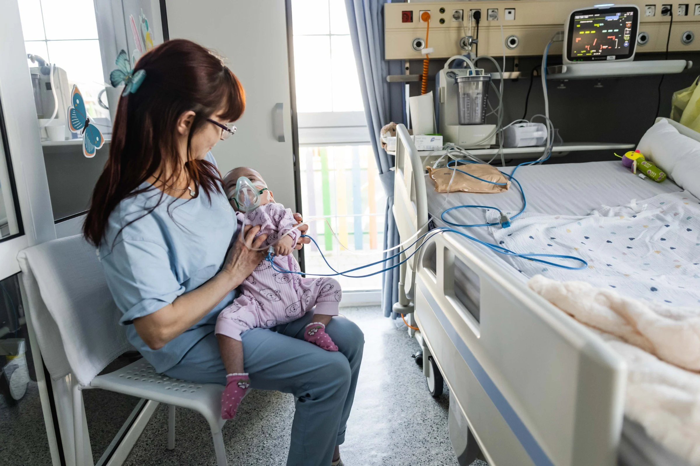 A woman in blue scrubs sits on a chair holding a baby in pink pajamas wearing a clear breathing mask held on by a strap, beside an empty hospital bed with tubing and a bedside monitor above it; a paper butterfly is stuck to the glass behind her
