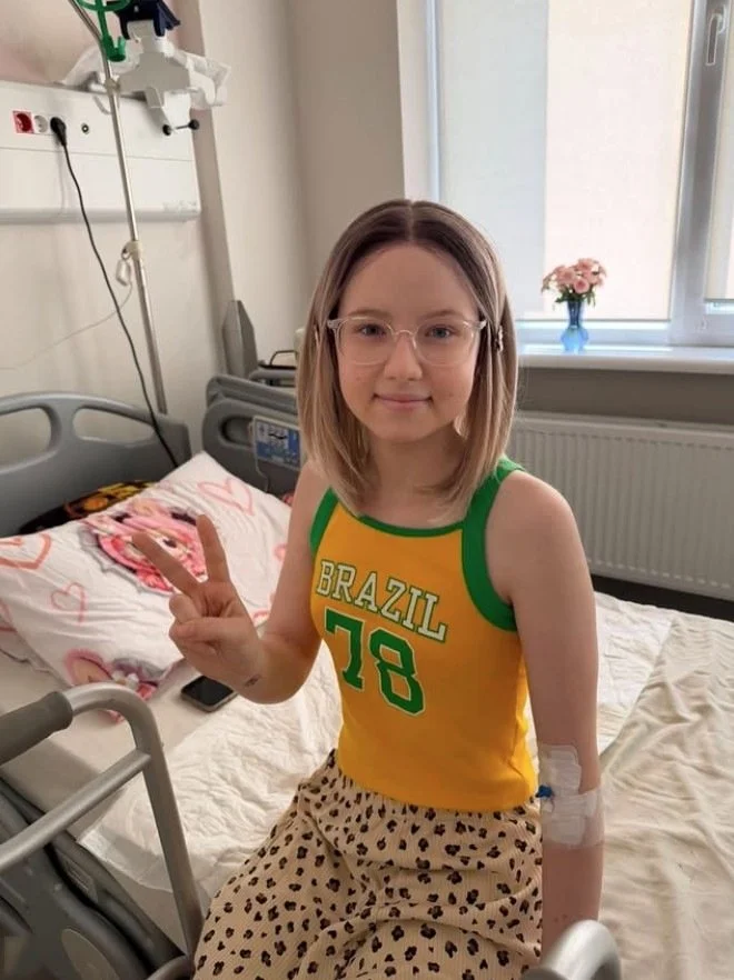 Yuliana sitting on the edge of a hospital bed by a window, wearing round glasses and a yellow and green tank top printed 'BRAZIL 78', holding up a peace sign, a walker in front of her and a small vase of pink roses on the windowsill