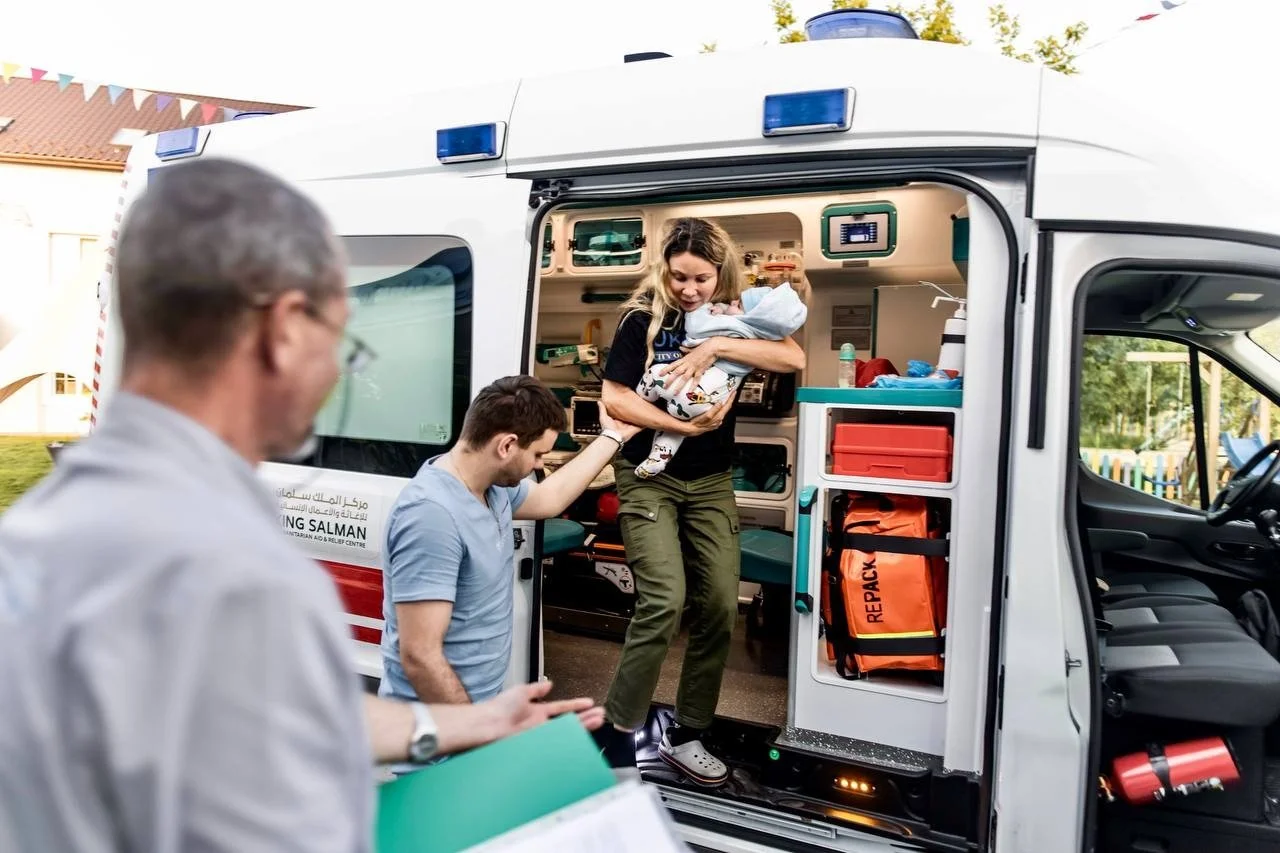 A woman carries a small child in her arms down from the open back of an ambulance, with two men standing beside it