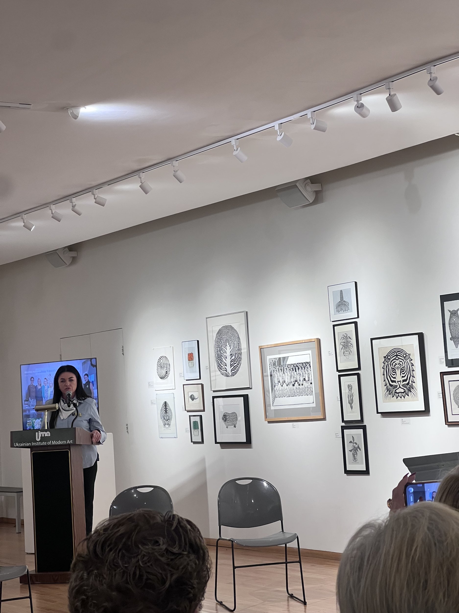 Oksana Taratula speaks into a microphone at the UIMA podium, with framed artworks behind her.